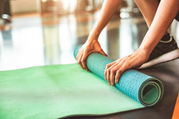 Close up of sporty woman folding yoga mattress in sport fitness gym training center background. Exercise mat rolling keeping after yoga class. Workout and sport training concept. Hands on carpet - Zdjęcie, obraz