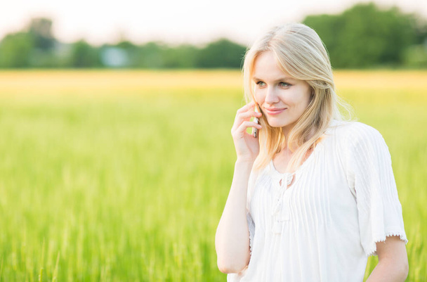 Mooie jonge vrouw spreken op mobiele telefoon tegen groene weide platteland zomer achtergrond. - Foto, afbeelding