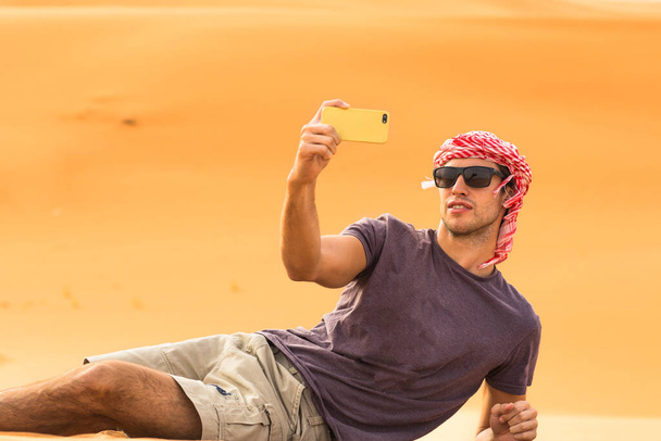 Utazás, turizmus és ifjúság. Fiatal férfi turista készít egy szelfi képet a mobiltelefonjával, miközben az Arab sivatag homokdűnéjén fekszik.. - Fotó, kép