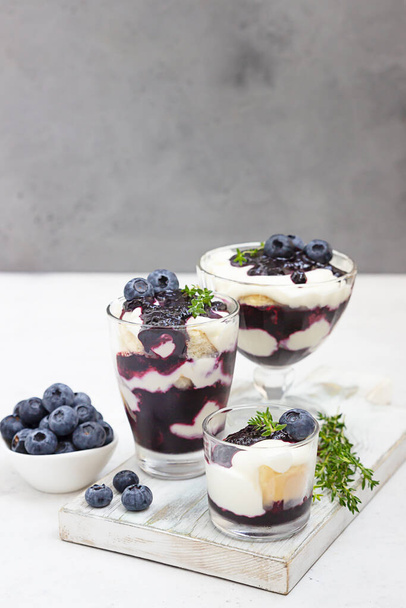Tiramisu. Homemade dessert in glasses with blueberries, cream and ladyfingers garnish with blueberries and thyme. Light grey background. - Foto, imagen