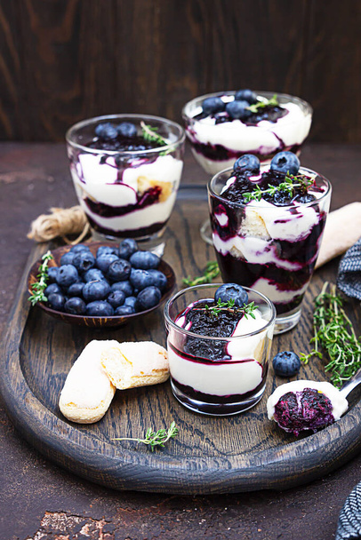 Dessert from mascarpone or ricotta cheese, blueberries and biscuit in glass on a dark concrete background. No baked cheesecake or tiramisu. - Fotoğraf, Görsel