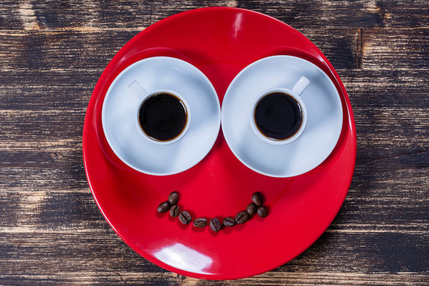 El plato rojo junto con dos tazas blancas de café y una sonrisa de granos de café en una mesa de madera. Primer plano, vista superior - Foto, imagen
