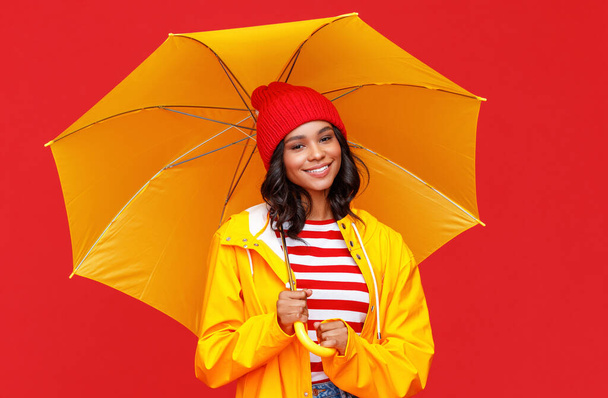 Positive ethnic female in hat and raincoat smiling and looking at camera while standing under umbrella on rainy day against red backgroun - Photo, Image