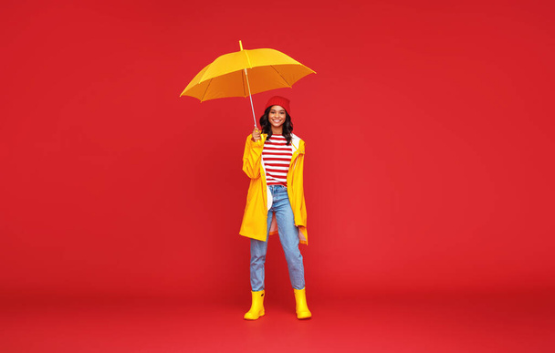 Volledige lengte optimistische etnische vrouw met paraplu glimlachen en kijken naar camera op regenachtige herfstdag tegen rode backgroun - Foto, afbeelding