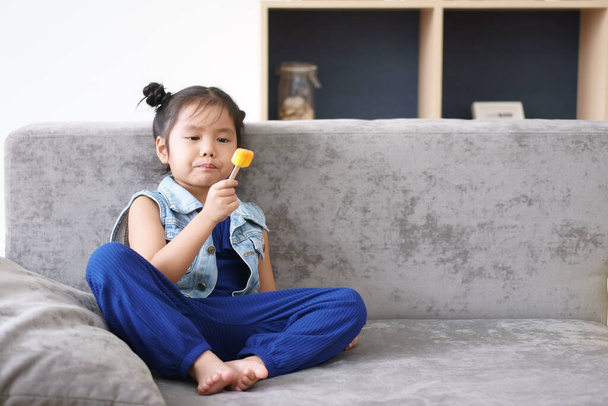 Azjatyckie dziecko słodkie lub dziewczynka szczęśliwa i cieszyć pyszne jedzenie pomarańczowe lody na słodki deser i odświeżyć na lato i kanapę w przedszkolu lub w domu przedszkola i przedszkola z jeansem nosić - Zdjęcie, obraz