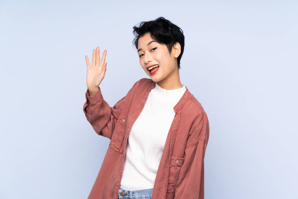 Jeune fille asiatique sur fond bleu isolé saluant avec la main avec expression heureuse - Photo, image