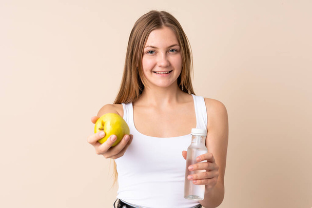 Teenager Ukrainian girl isolated on beige background with an apple and with a bottle of water - Foto, immagini