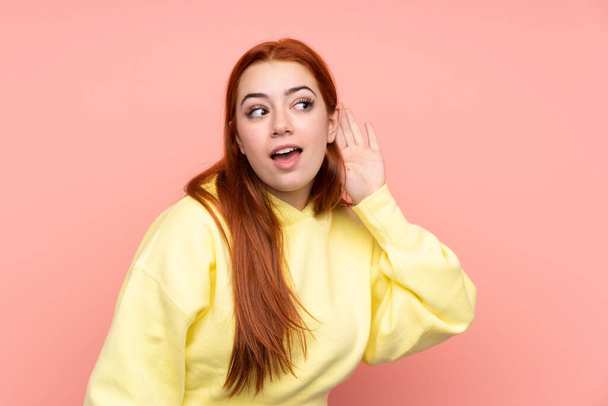 Redhead teenager girl over isolated pink background listening to something by putting hand on the ear - Foto, Imagem