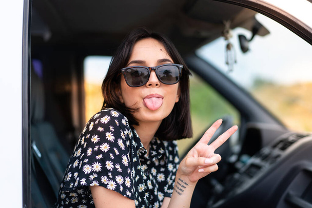 Joven mujer caucásica sonriendo y mostrando signo de victoria con una cara alegre en una furgoneta al aire libre - Foto, Imagen