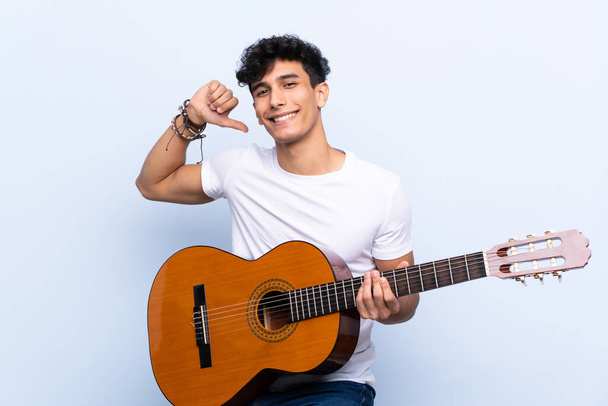 Młody argentyński człowiek z gitarą na odizolowanym tle niebieski dumny i zadowolony z siebie - Zdjęcie, obraz