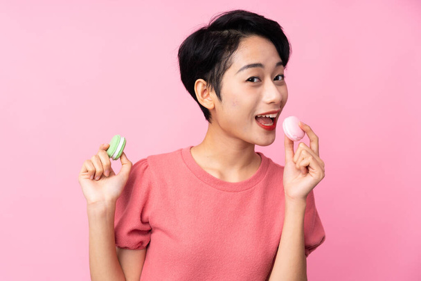 Joven asiática chica sobre aislado rosa fondo celebración colorido francés macarons y comer ella - Foto, imagen
