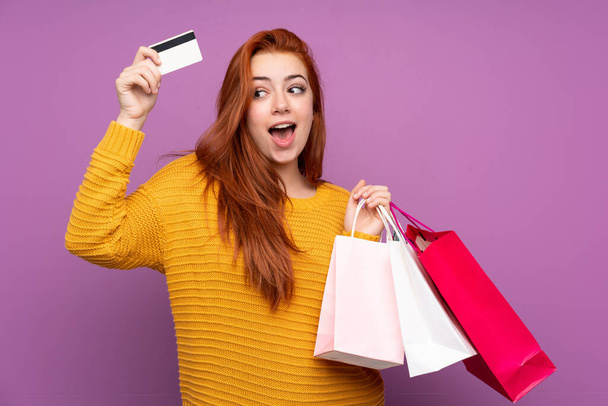 Redhead teenager girl over isolated purple background holding shopping bags and a credit card - Zdjęcie, obraz