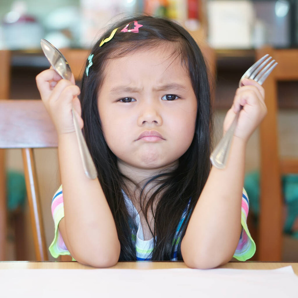 Azjatyckie dziecko głodny lub dziecko dziewczyna gospodarstwa łyżka z widelcem i nieszczęśliwy lub zły i drażliwy grymas przez długi czas czekać jeść pyszne jedzenie lub anoreksja i nudne jedzenie na śniadanie rano w restauracji - Zdjęcie, obraz