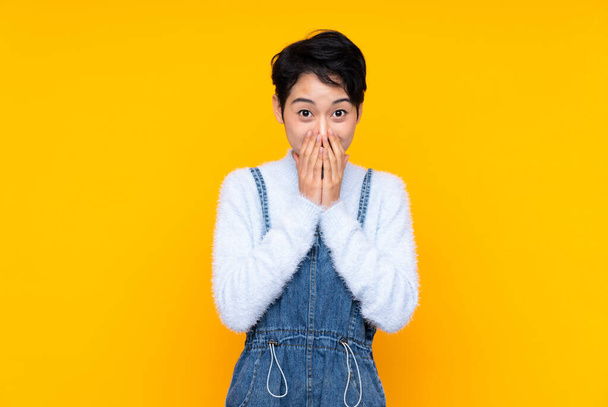 Young Asian girl in overalls over isolated yellow background with surprise facial expression - Photo, Image