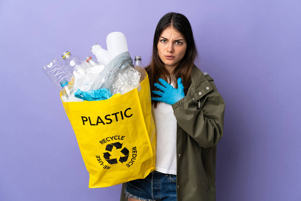 Junge Frau mit einer Tüte voller Plastikflaschen zum Recyceln isoliert auf violettem Hintergrund überrascht und schockiert, während sie nach rechts schaut - Foto, Bild