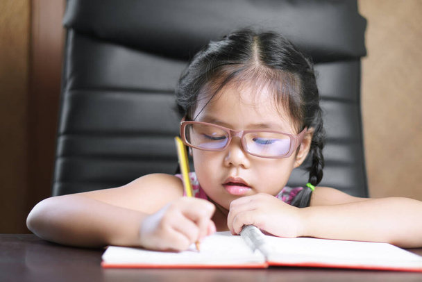 ázsiai gyermek vagy gyerek lány visel szemüveg rövidlátó és szorgalmas csinál házi feladat vagy tanulás írás fehér papír könyv és íróasztal vagy asztal óvodai óvoda vagy óvoda iroda - Fotó, kép