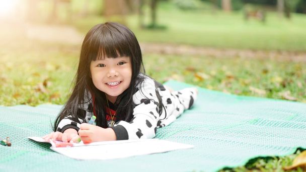 Niño asiático lindo o niña aprendiendo para colorear o dormir dibujo pintura en estera verde y prado de la naturaleza en otoño jardín de invierno con sonrisa feliz dientes blancos y educación preescolar en vacaciones relajarse - Foto, imagen