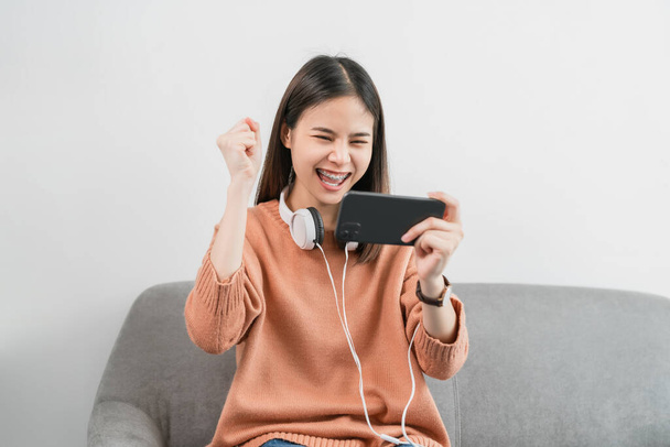 Excited young Asian woman wear white headphone on the head and playing games on smartphone in the living room at home. - Photo, Image