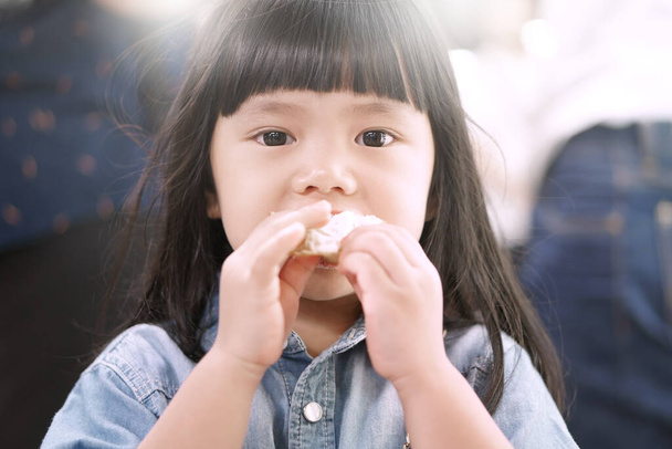 Asijské dítě roztomilé nebo dítě dívka a dlouhé vlasy hladové a vychutnat jíst chléb jídlo jako dezert nebo svačinka a nosit džíny na ulici a příroda slunce - Fotografie, Obrázek