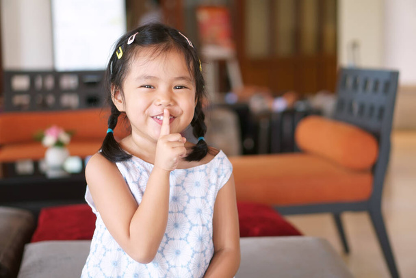Asian child cute or kid girl smile with laugh squat and forefinger close mouth for tell quietly shh or stop silence and secret surprise with play hide and seek at library at school or reception room - Photo, Image