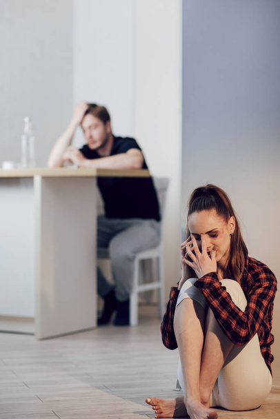 Victim of domestic violence. Young scared woman with bruises on face complains to girlfriend on cell phone sitting on the floor in the kitchen. Her husband is sitting behind and drinking alcohol - Fotoğraf, Görsel