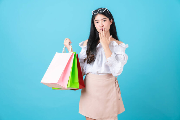 Joven mujer asiática sonriente sosteniendo bolsas de compras multicolores y manos a la boca sobre fondo azul claro. - Foto, Imagen