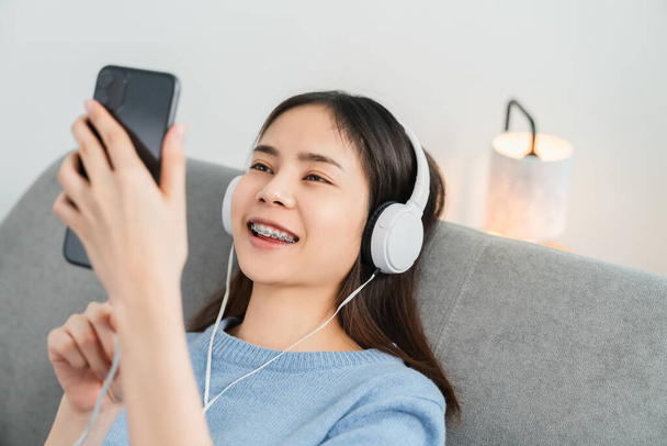 Mutlu gülümseyen Asyalı kadın beyaz kulaklıklardan müzik dinliyor ve evde tatilde akıllı telefonlara dokunuyor.. - Fotoğraf, Görsel