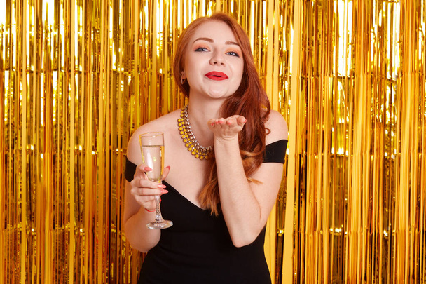 Excited pretty red haired girl holding wineglass of sparkling champagne standing isolated on sparkle background. Model sending air kisses while celebrating important event. - Фото, изображение