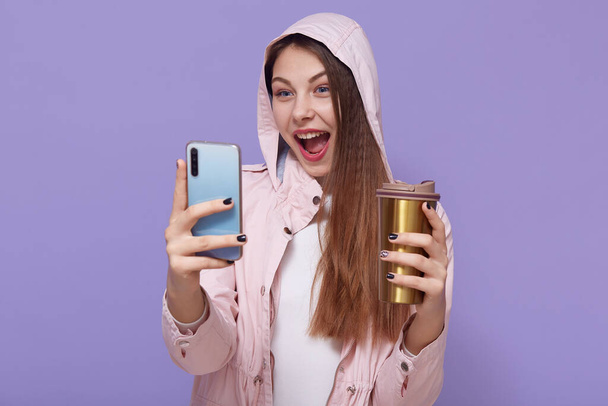Attractive Caucasian girl making selfie against lilac background, being excited, keeping mouth widely opened, enjoying hot beverage, wearing pale pink raincoat. - Φωτογραφία, εικόνα