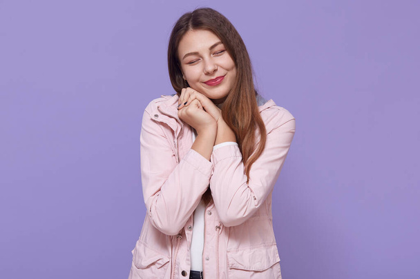 Pretty smiling charming young woman wearing pale pink jacket holding fists near cheeks isolated on lilac wall, keeping eyes closed, looks cute, female with long beautiful hair. - Photo, image