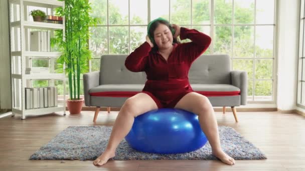 Gros asiatique femme exercice sur gymnase ballon tandis que heureux port casque écoute de la musique. - Séquence, vidéo