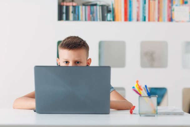 Młody chłopak siedzi przy stole z laptopem i przygotowuje się do szkoły. Koncepcja edukacji internetowej. - Zdjęcie, obraz