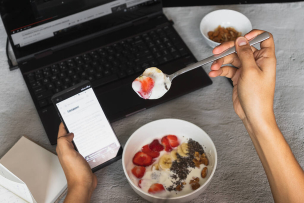A high angle shot of a person eating yogurt with fruits while using his phone - Valokuva, kuva
