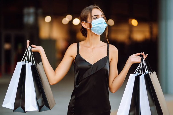 Young woman  in protective sterile medical mask on her face with shopping bags enjoying in shopping. Purchases, black friday, sale concept. Covid- 2019. - 写真・画像