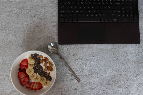 Μια κορυφαία άποψη ενός μπολ γιαούρτι με φρούτα στο τραπέζι με ένα φορητό υπολογιστή σε αυτό - Φωτογραφία, εικόνα