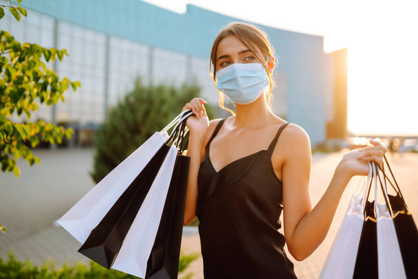 Fashionable woman wearing protective medical mask with shopping bags at sunset. Purchases, black friday, sale concept. Covid- 2019. - Foto, afbeelding
