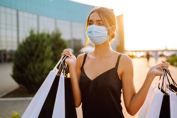 Fashionable woman wearing protective medical mask with shopping bags at sunset. Purchases, black friday, sale concept. Covid- 2019. - Foto, Imagem