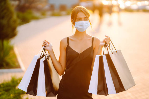 Fashionable woman wearing protective medical mask with shopping bags at sunset. Purchases, black friday, sale concept. Covid- 2019. - 写真・画像