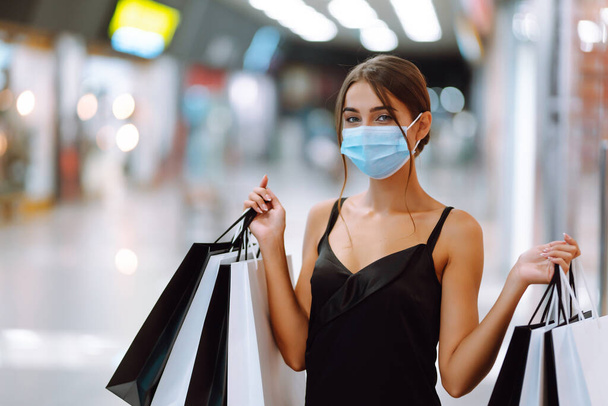 Woman in shopping. Young woman in protective sterile medical mask on her face with shopping bags in the mall. Purchases, black friday, discounts,  sale concept. Covid-2019. - Фото, изображение
