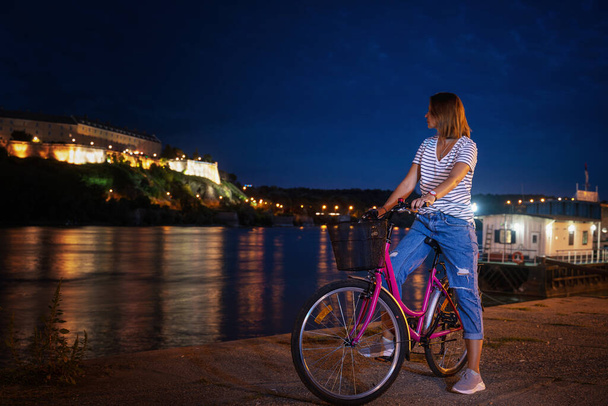 Young woman with a bike on the Novi Sad embankment overlooking the Petrovaradin fortress, night shot, travel to Serbia, Balkan countries - Fotoğraf, Görsel