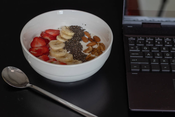 A closeup of a bowl of yogurt with fruits on the table with a laptop on it - Φωτογραφία, εικόνα