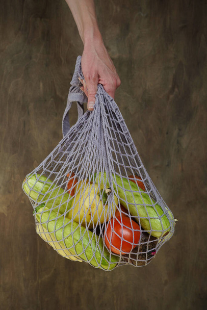 Woman holds her hand gray bag string bag with vegetables,zucchini,bell peppers,carrots,beets,onions brown wooden background.Harvest from garden fresh bright vegetables.Concept zero waste. - Foto, Bild