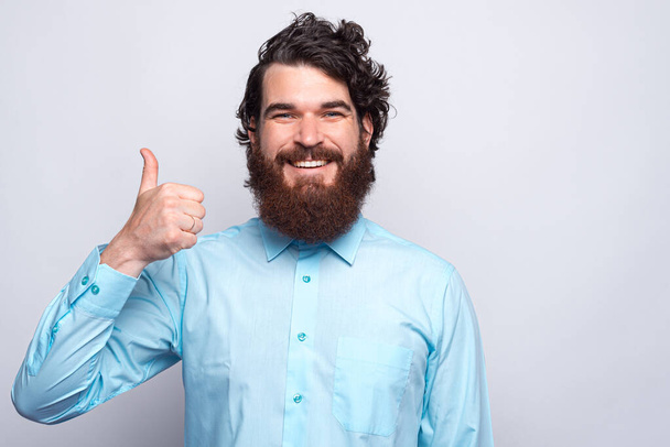 Portret van vrolijke jonge man met baard in blauw shirt met duim omhoog en glimlach - Foto, afbeelding