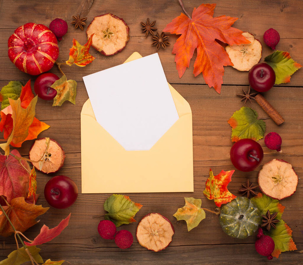 Autumn floral composition with pumpkins and empty blank card in paper envelope. Wreath of dry maple leaves, flowers and apple. Wooden rustic table background. Fall, Halloween or Thanksgiving Holiday design. Flat lay, top view. - Photo, Image