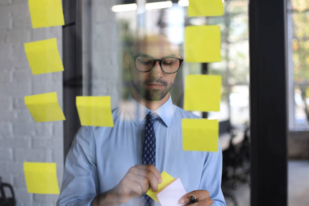 Young modern business man using adhesive notes while standing behind the glass wall in the office - Photo, image