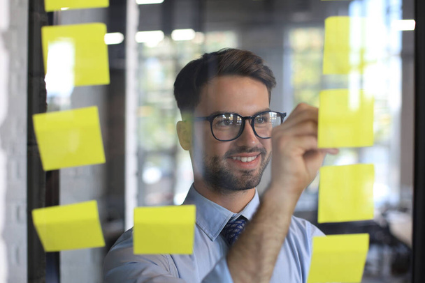 Young modern business man using adhesive notes while standing behind the glass wall in the office - Photo, Image