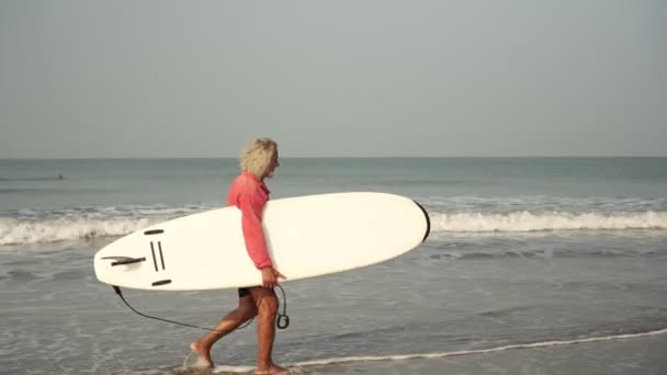Een oude man loopt langs het strand met een surfplank. Een actieve gepensioneerde gaat sporten. Opa is een surfer. - Video