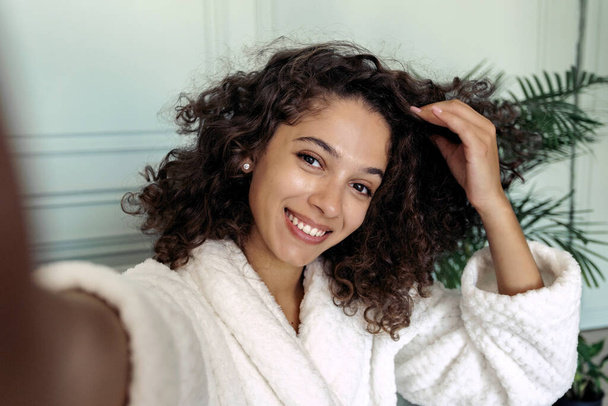 Ritratto di giovane donna afro americana felice in accappatoio scattarsi foto sulla macchina fotografica, trascorrere il fine settimana a casa, seduta in soggiorno, fare streaming online per il blog di bellezza - Foto, immagini