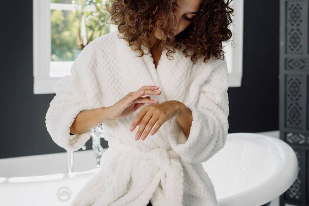 Concept van hydraterende procedure en dagelijkse routine. Afrikaans-Amerikaanse vrouw aanbrengen crème op handen, staan in badjas, het doorbrengen van de ochtend in de badkamer - Foto, afbeelding