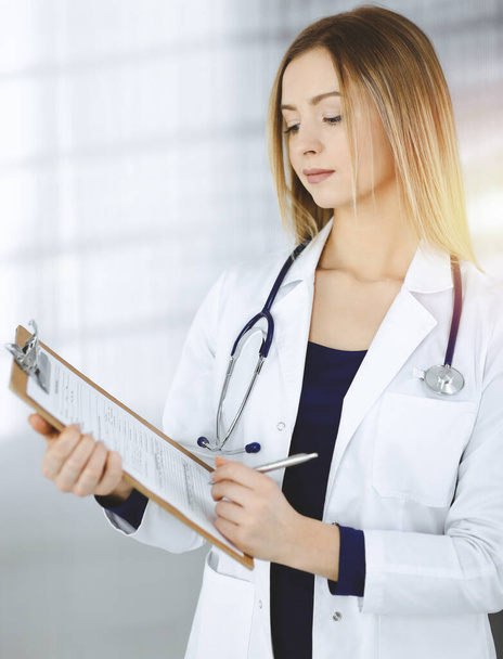 Young optimistic woman-doctor is holding a clipboard in her hands, while standing in a sunny clinic. Portrait of friendly female physician with a stethoscope. Perfect medical service in a hospital - Foto, Imagem